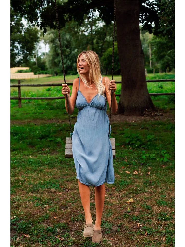 A woman in a Germaine des Prés Josephine Grey Nightie swings on a wooden swing in a grassy outdoor area with trees and a fence in the background.