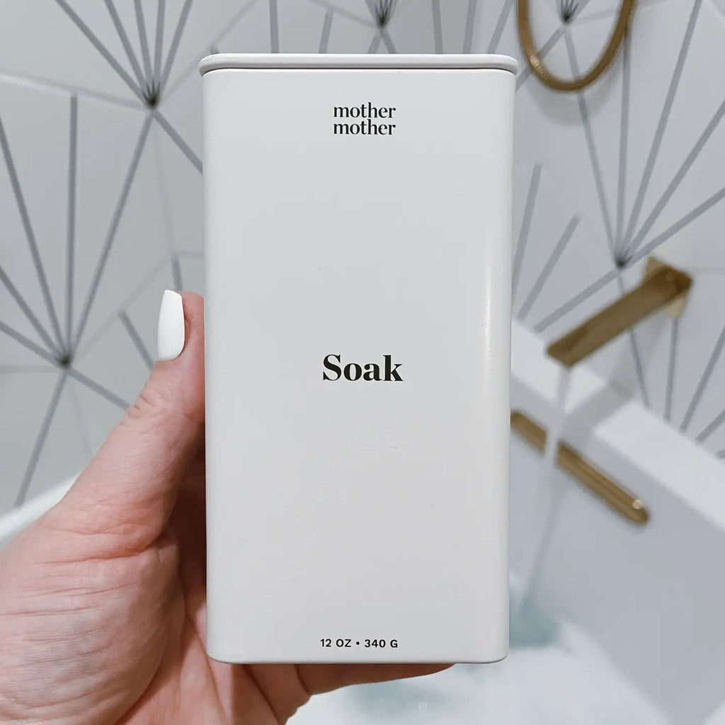 A hand holds a rectangular white container labeled "Mother Mother Magnesium Bath Soak" with geometric tiles and brass hardware in the background, suggesting a magnesium soak infused with lavender essential oil.