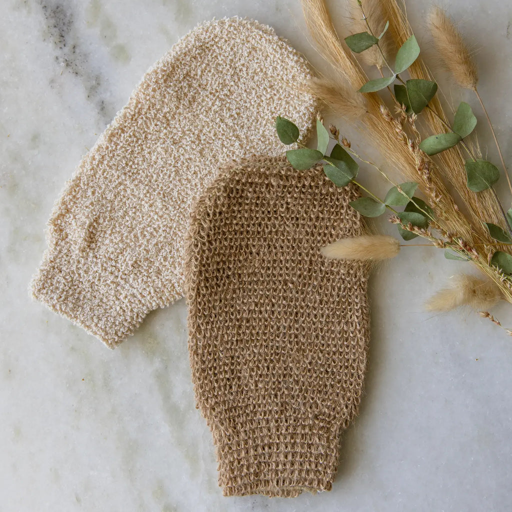 The Earth Ahead Ramie Bath Shower Mitts, one in cream and the other in brown, rest on a marble surface beside a small bouquet of dried plants.