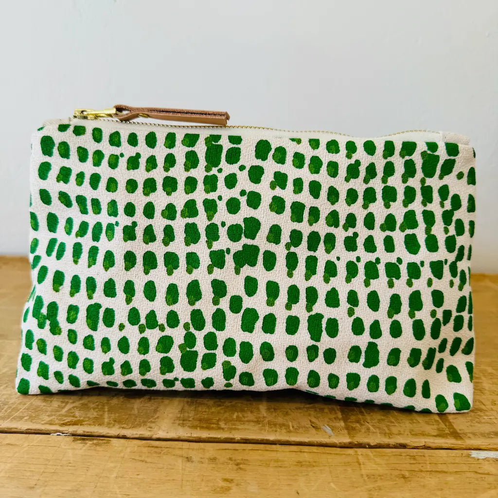 White fabric pouch with a green abstract dot pattern and a zipper, placed on a wooden surface.
