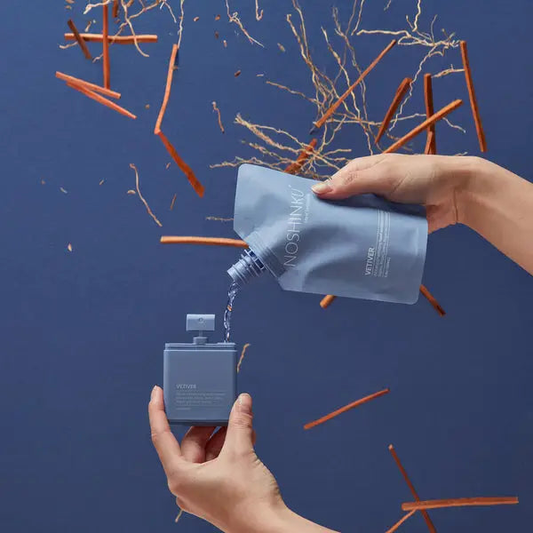 A person pouring a liquid from a flexible pouch into a small, open container against a blue background with plant twigs and shavings scattered around.
