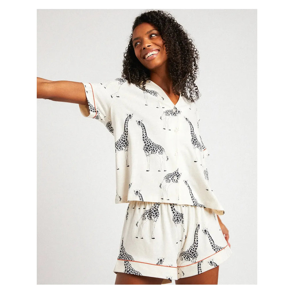 A person with curly hair smiles and poses in a Chelsea Peers Organic Cotton Giraffe Print pajama set made from soft organic cotton.