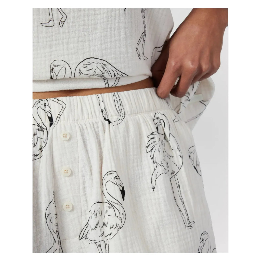 Close-up of a person adjusting the waistband of white shorts with a black flamingo print design, made from lightweight cotton cheesecloth, showing a matching top partially visible. The product is the Chelsea Peers Cotton Cheesecloth Flamingo Sketch Print Cami Short Pajama Set.