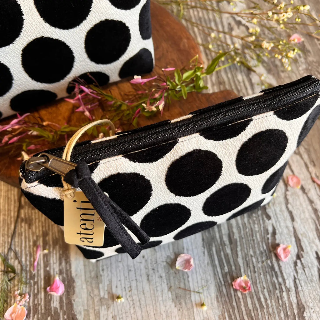 A polka-dotted pouch with a zipper and a branded tag rests on a wooden surface among decorative flowers.