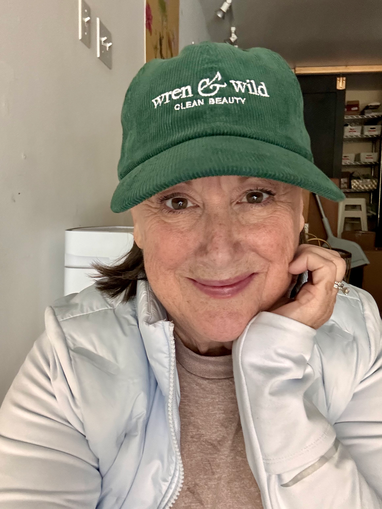 A person sporting a Wren and Wild Green Corduroy Logo Hat and a light jacket smiles at the camera indoors, exuding effortless swag.