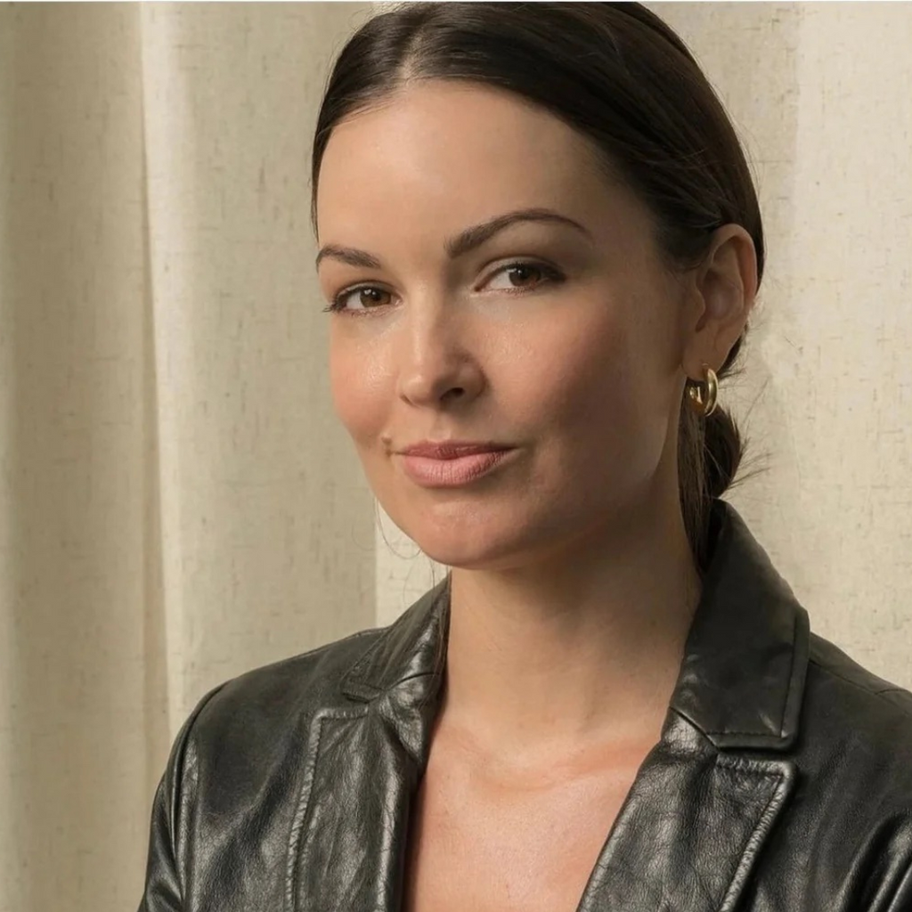 A woman with dark hair tied back, wearing a black leather jacket and gold hoop earrings, stands against a light-colored curtain background, as if ready for her Berlin Skin Matcha Mini Facials tour or perhaps after one of those rejuvenating mini facials.