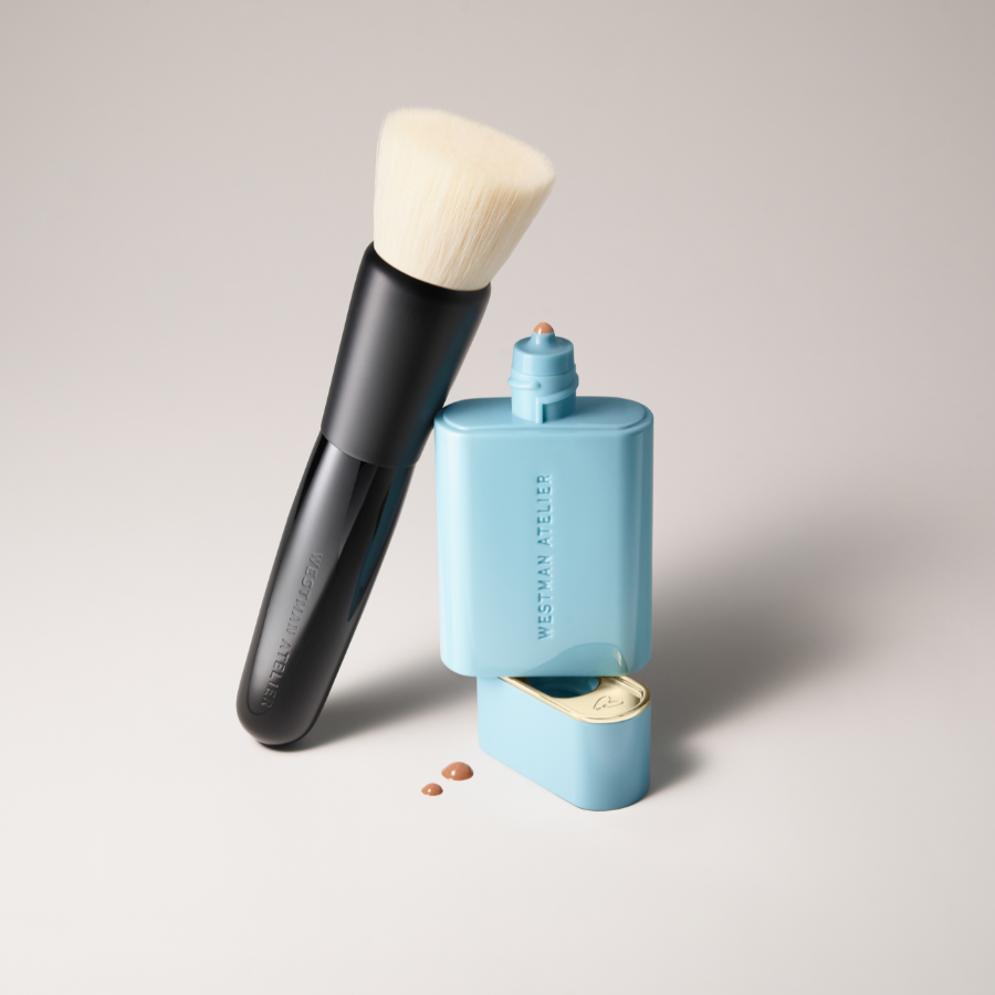 Experience the elegance of Westman Atelier at The Makeup Event, March 6-8. Enhance your glow with their Soleil Parfait liquid bronzer, perfectly complementing a minimalist scene featuring a makeup brush and blue foundation container accented by spilled drops.