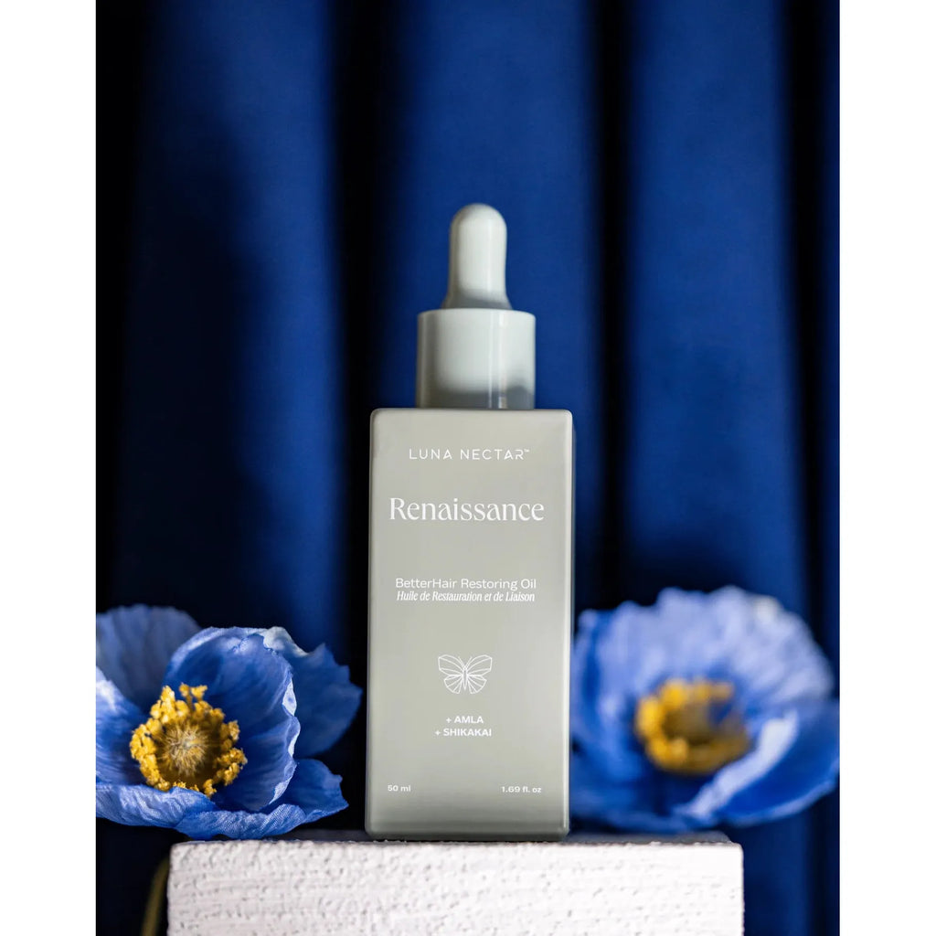 A bottle of luna nectar renaissance hair restoring oil displayed with blue flowers against a dark blue backdrop.