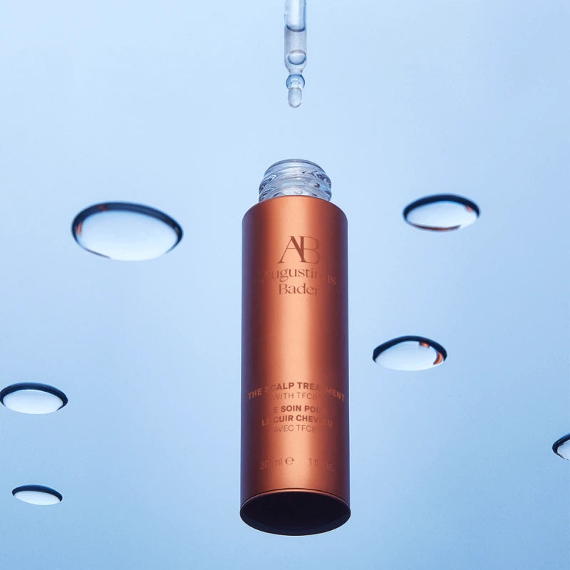 A bottle of augustinus bader scalp treatment suspended with droplets of liquid falling around it against a blue background.