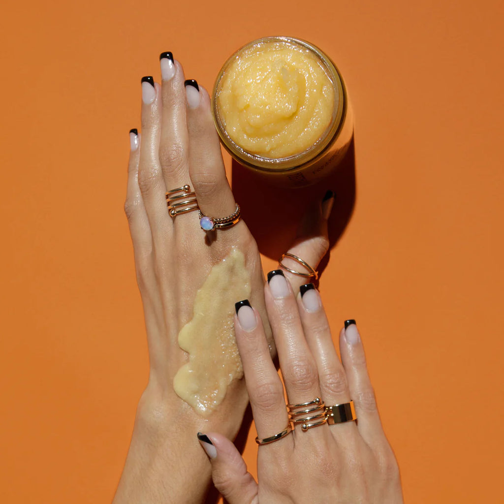 A person showcasing their hands with black-tipped manicure and multiple rings, holding a jar of cosmetic cream with some of the product on their hand against an orange background.