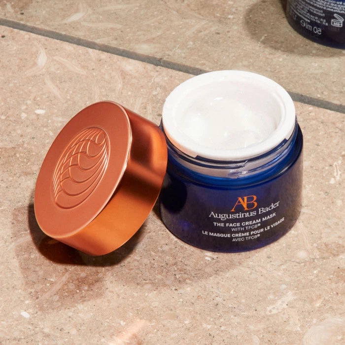 An opened jar of augustinus bader face cream on a tiled surface.