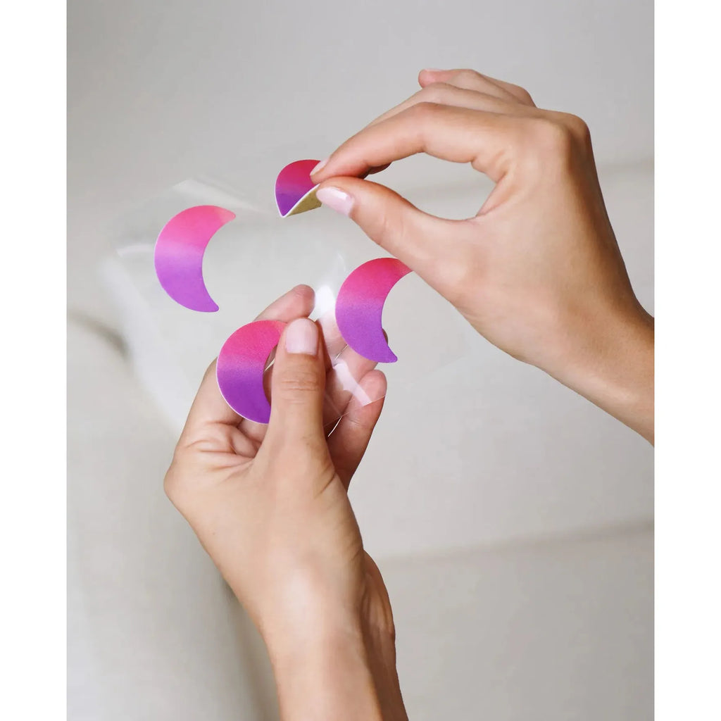 A person is peeling a purple crescent-shaped sticker off a clear backing with their fingers.
