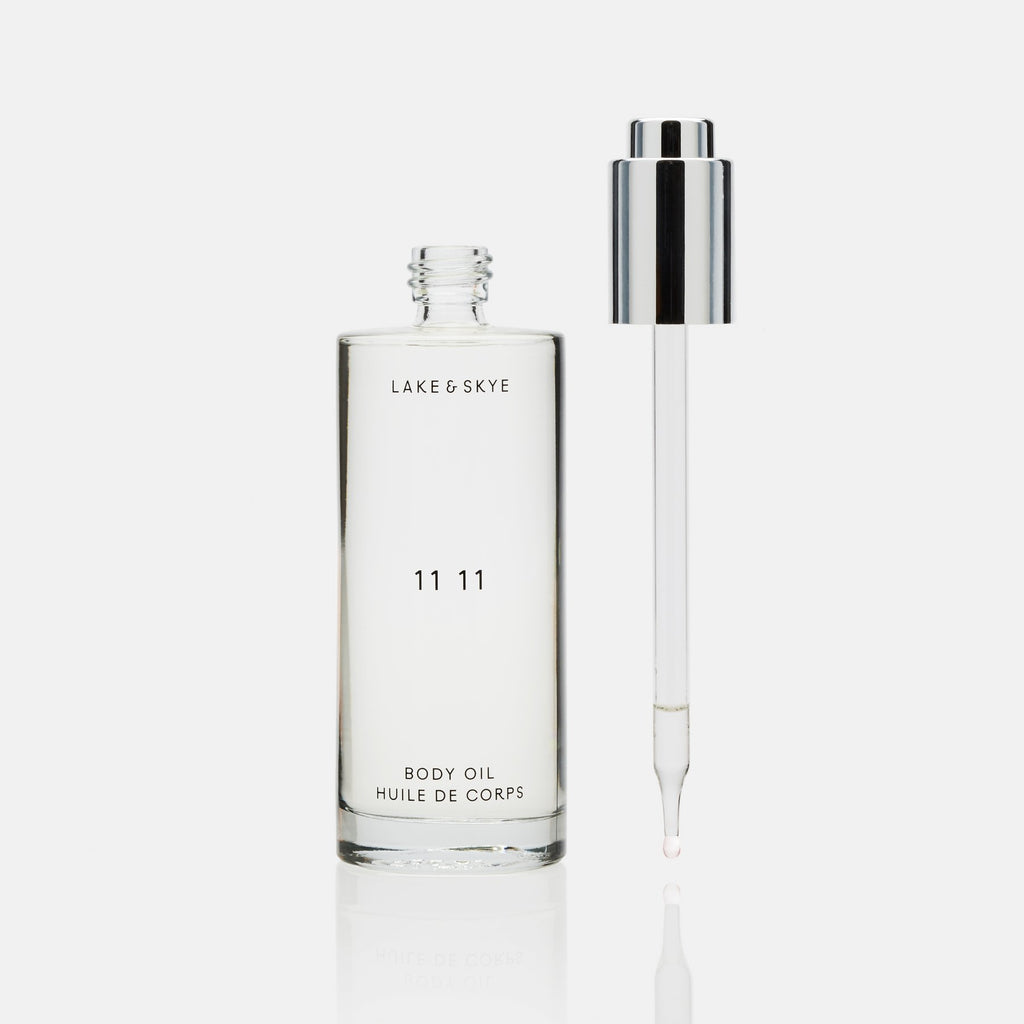 A transparent bottle of lake & skye '11 11' body oil with a silver dropper, some oil visible on the dropper tip, isolated against a white background.