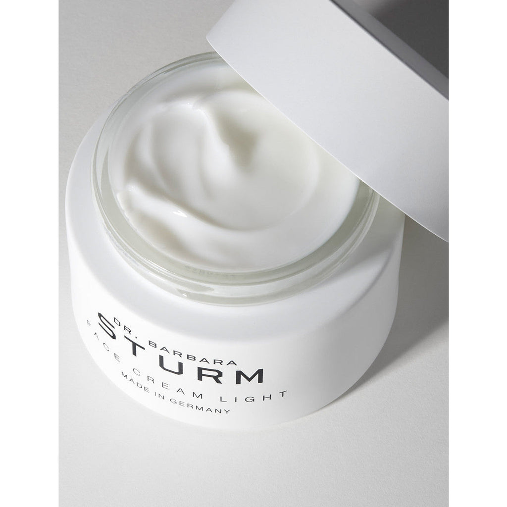 A jar of barbara sturm face cream light on a white surface with its lid partially open, showcasing the product inside.