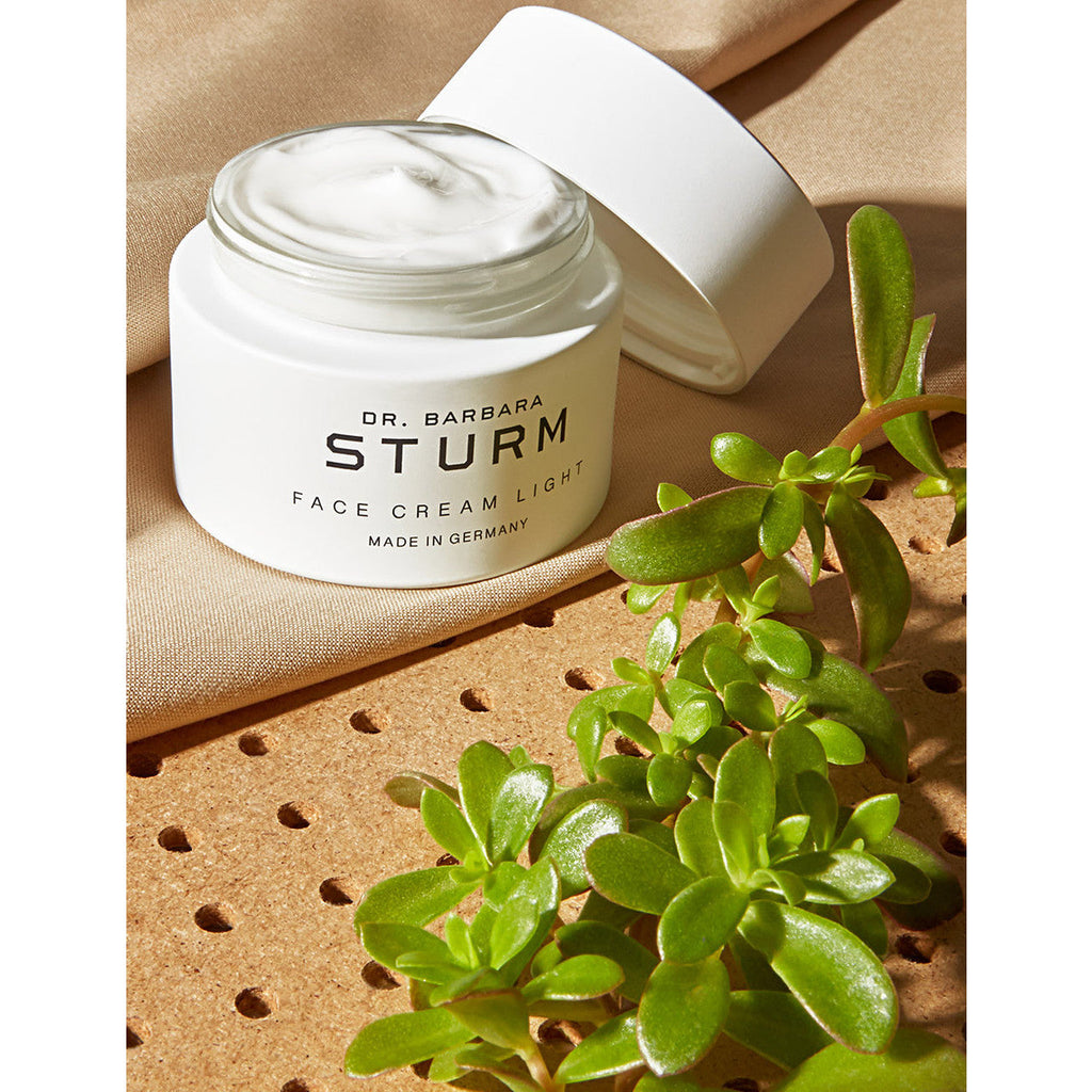 A jar of dr. barbara sturm face cream light placed on a textured surface alongside a green succulent plant.