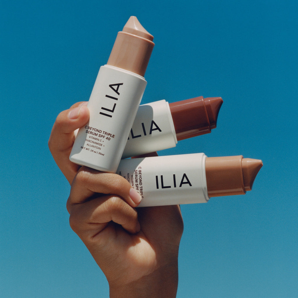 A hand holding three tubes of ilia brand cosmetics against a blue background.