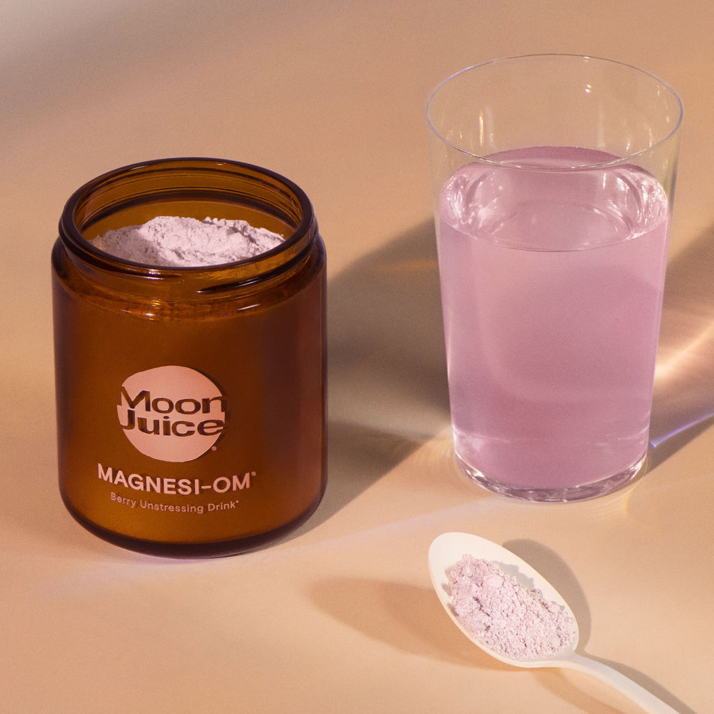 A jar of moon juice magnesi-om supplement with a glass of mixed pink beverage and a scoop of powder beside it.