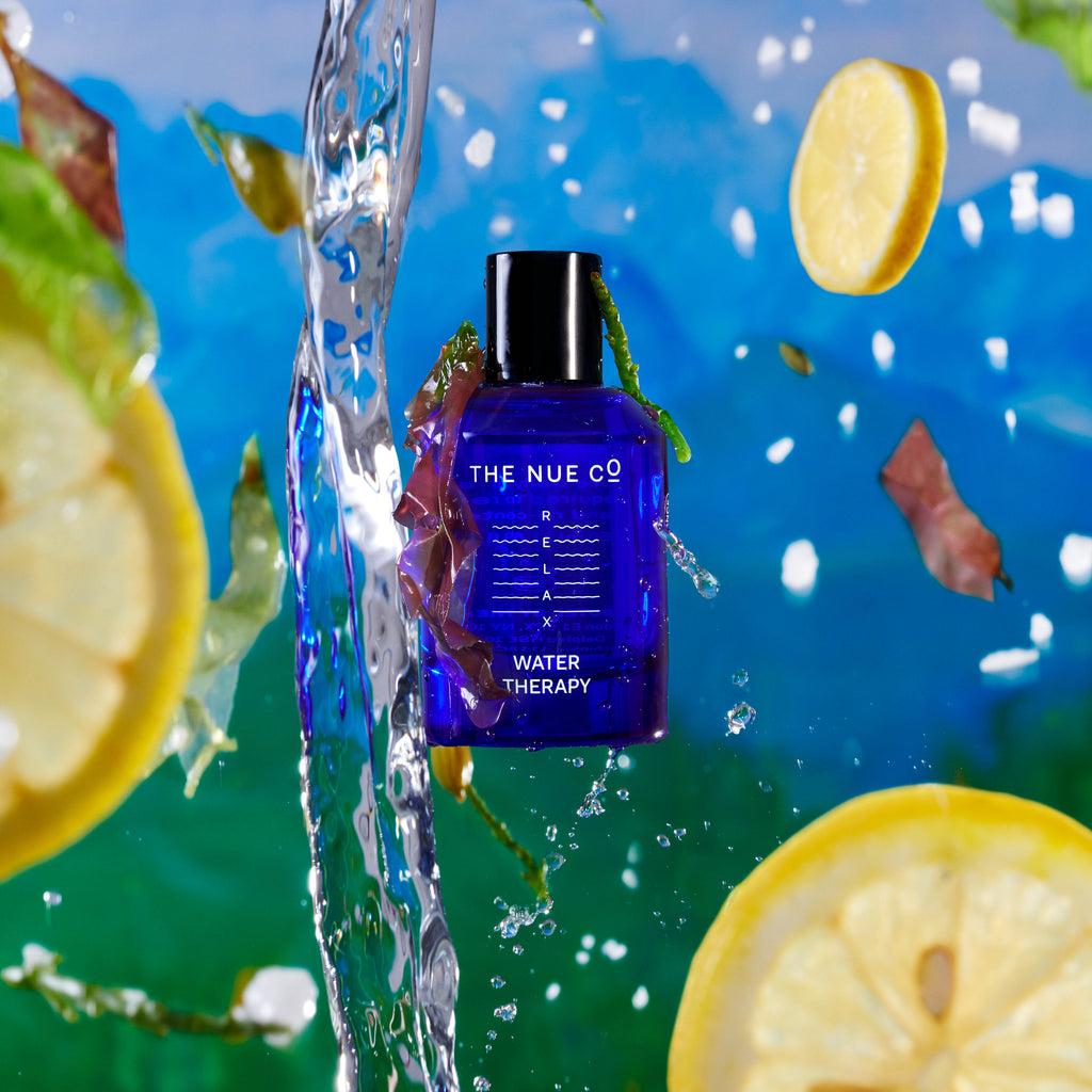 Product showcase of "the nue co. water therapy" bottle with water splashing and lemon slices against a blue background.