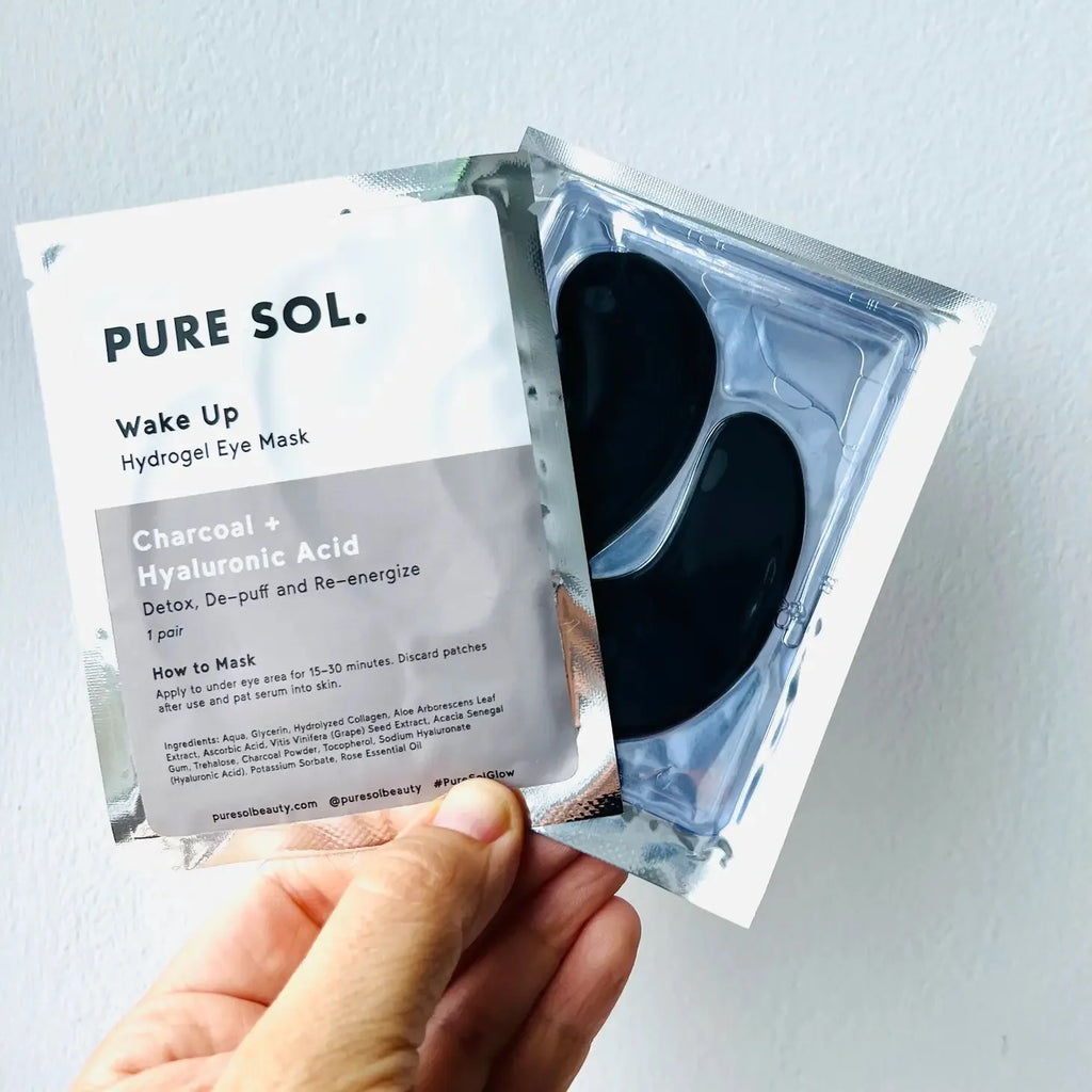 A hand holding a package of "pure sol. wake up hydratedgel eye mask" with charcoal and hyaluronic acid.