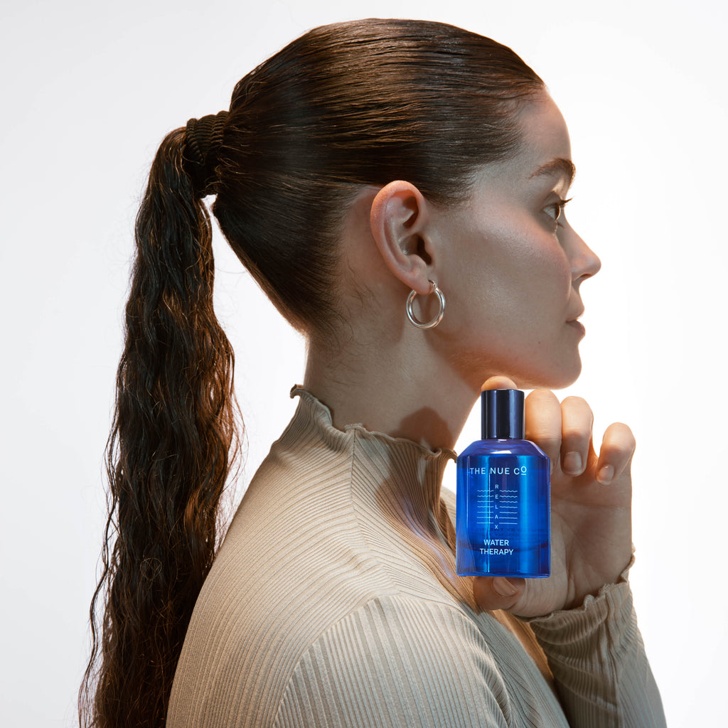 A woman holding a blue bottle of skincare product near her face with a neutral background.