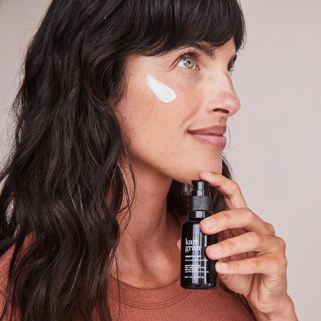 Woman with a dab of cream on her cheek holding a skincare product bottle.