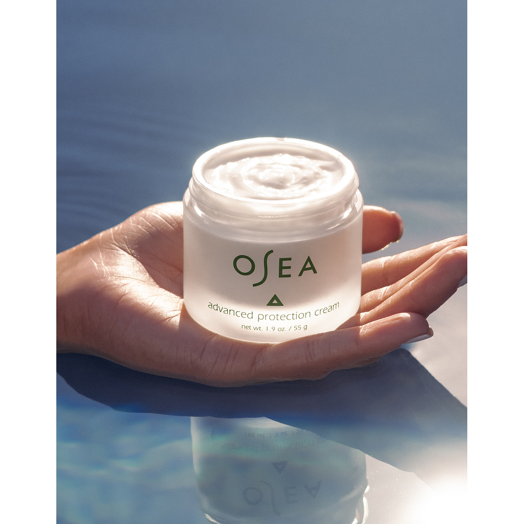 A hand holding a jar of osea advanced protection cream against a tranquil blue water background.