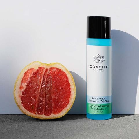 A bottle of odacite blue aura cleansing water next to a sliced grapefruit on a neutral background.