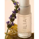 A bottle of ilia beauty product with a sprig of lavender against a two-tone background.