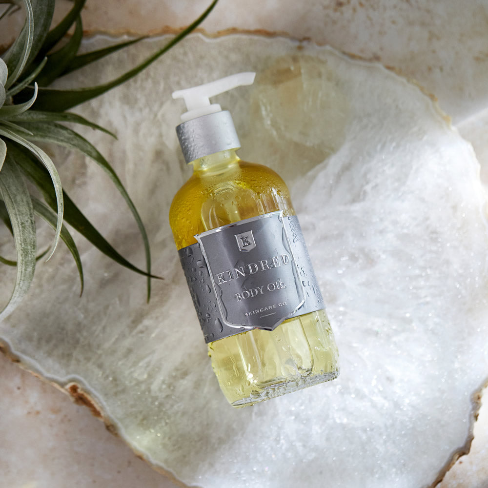 Bottle of body oil on a natural stone dish with a plant beside it.