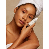 Woman holding a skincare product to her cheek, wrapped in a towel with her hair up.