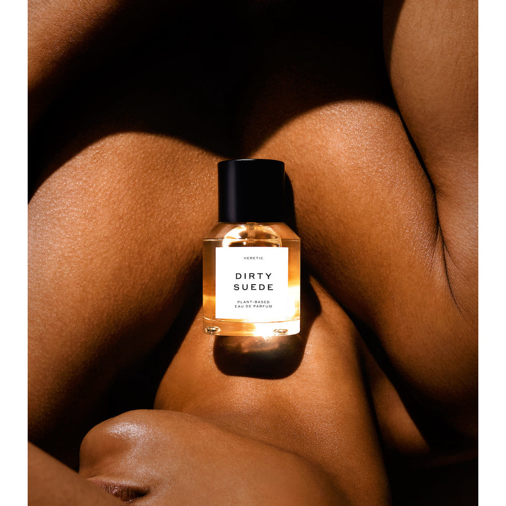 A bottle of perfume with the label "dirty suede" placed between folds of smooth, dark skin bathed in warm light.