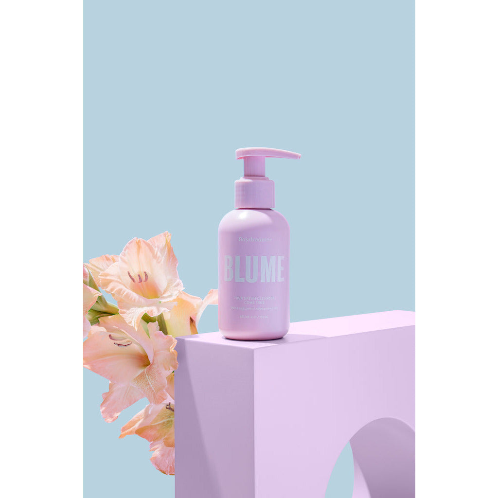 A bottle of blume skincare lotion next to a flower on a pastel background.