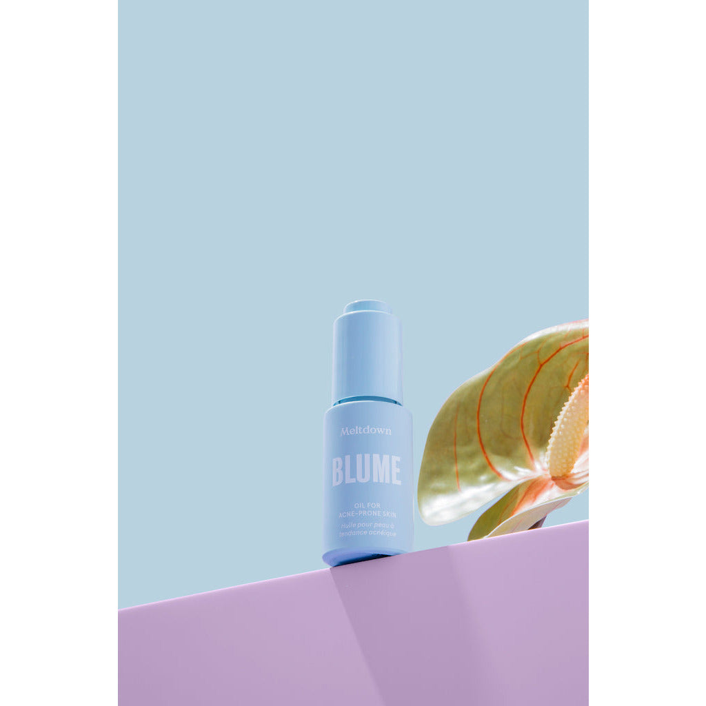 A bottle of blume meltdown acne treatment oil placed against a two-tone background with a plant leaf partially in view.