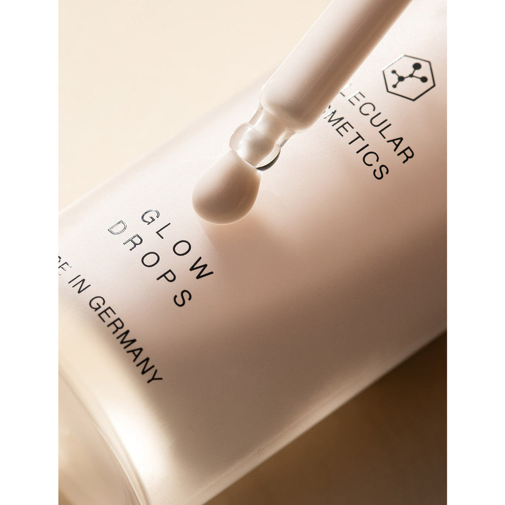 A dropper dispenses a liquid cosmetic product labeled "glow drops" onto a surface.