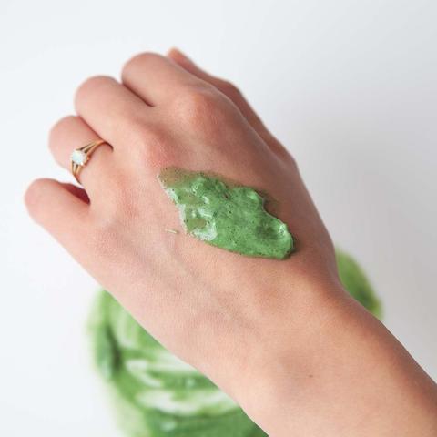 A hand with a gold ring, testing a dab of green cosmetic face mask on the back of the hand.