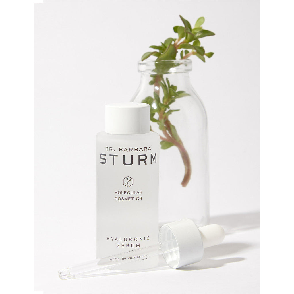 A bottle of dr. barbara sturm hyaluronic serum with a pipette and green plant in the background.