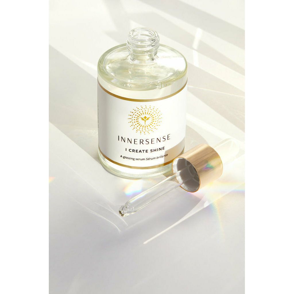 A bottle of innersense "i create shine" serum next to a dropper with sunlight casting shadows and reflections on a white surface.