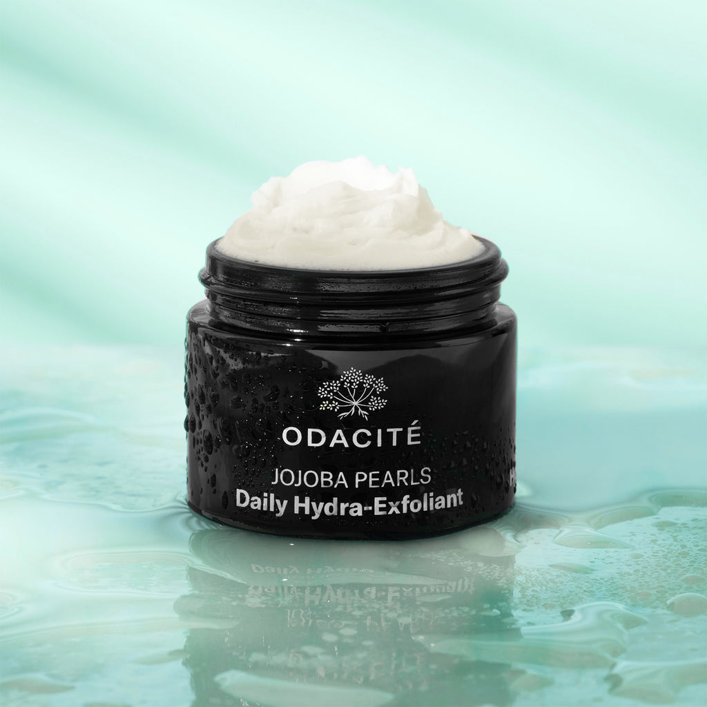 Open jar of odacite jojoba pearls daily hydra-exfoliant cream on a reflective surface with water droplets.