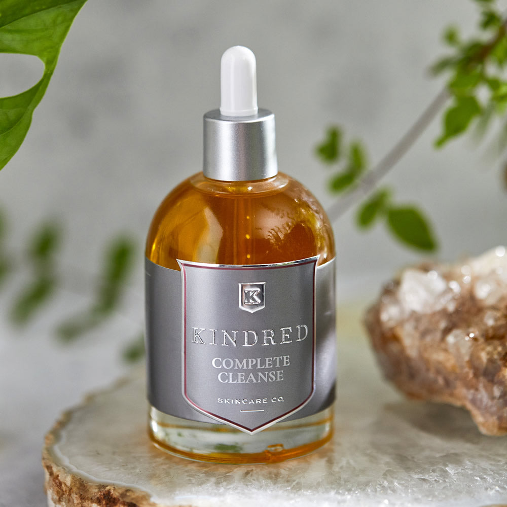 A bottle of kindred complete cleanse skincare oil with a dropper, displayed on a natural stone surface with greenery and a crystal in the background.