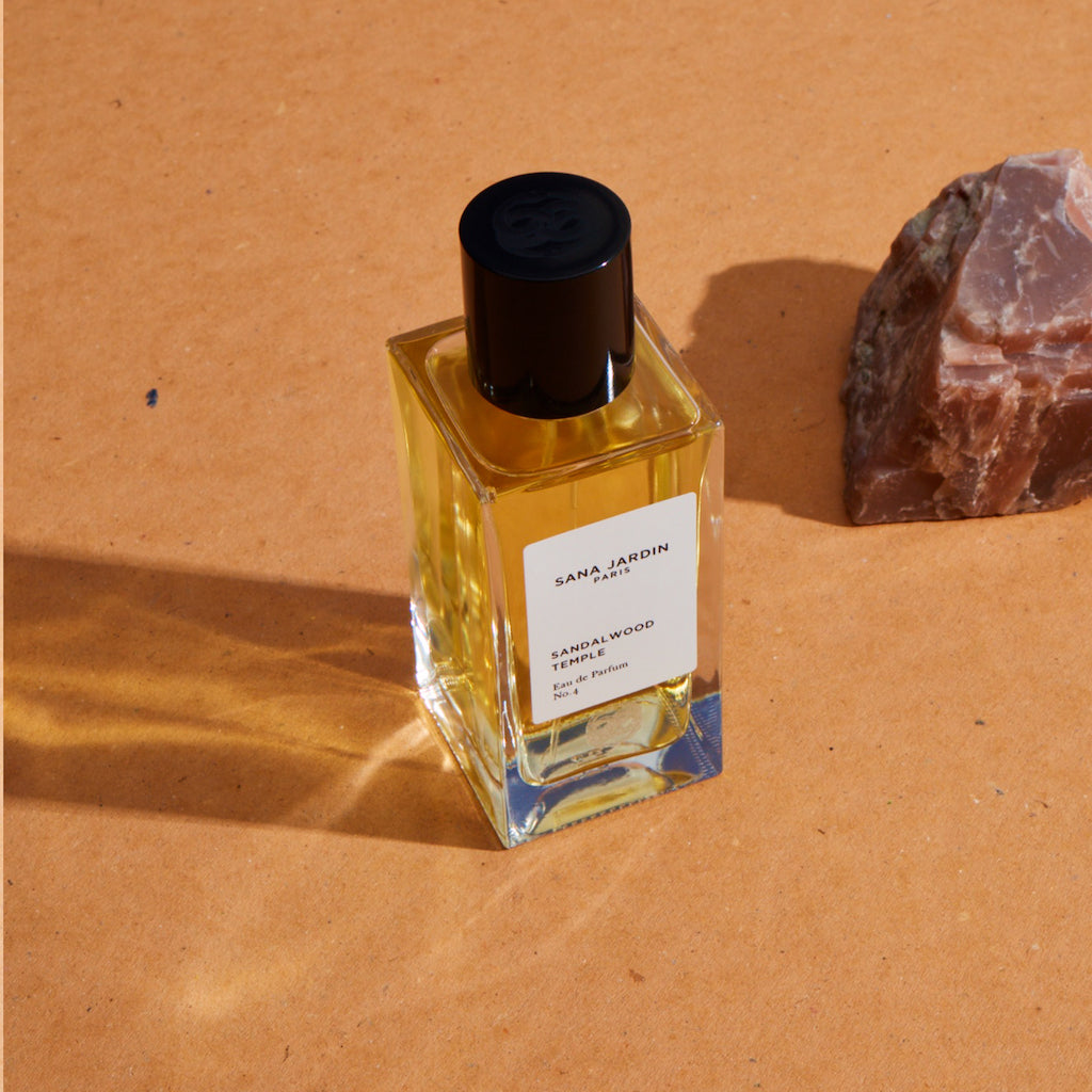 Glass perfume bottle labeled "santalwood" beside a piece of stone on an earth-toned surface.