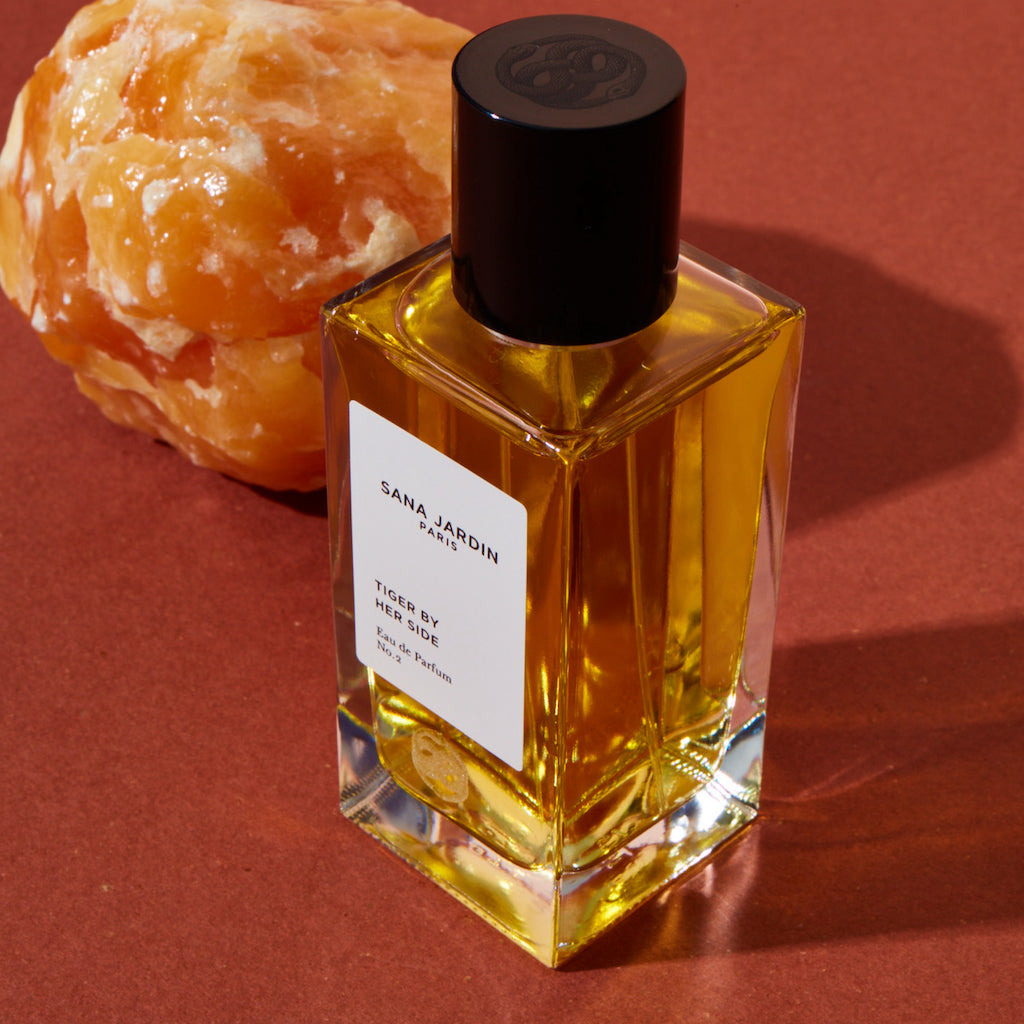 A bottle of sana jardin perfume next to a peeled citrus fruit on a maroon background.