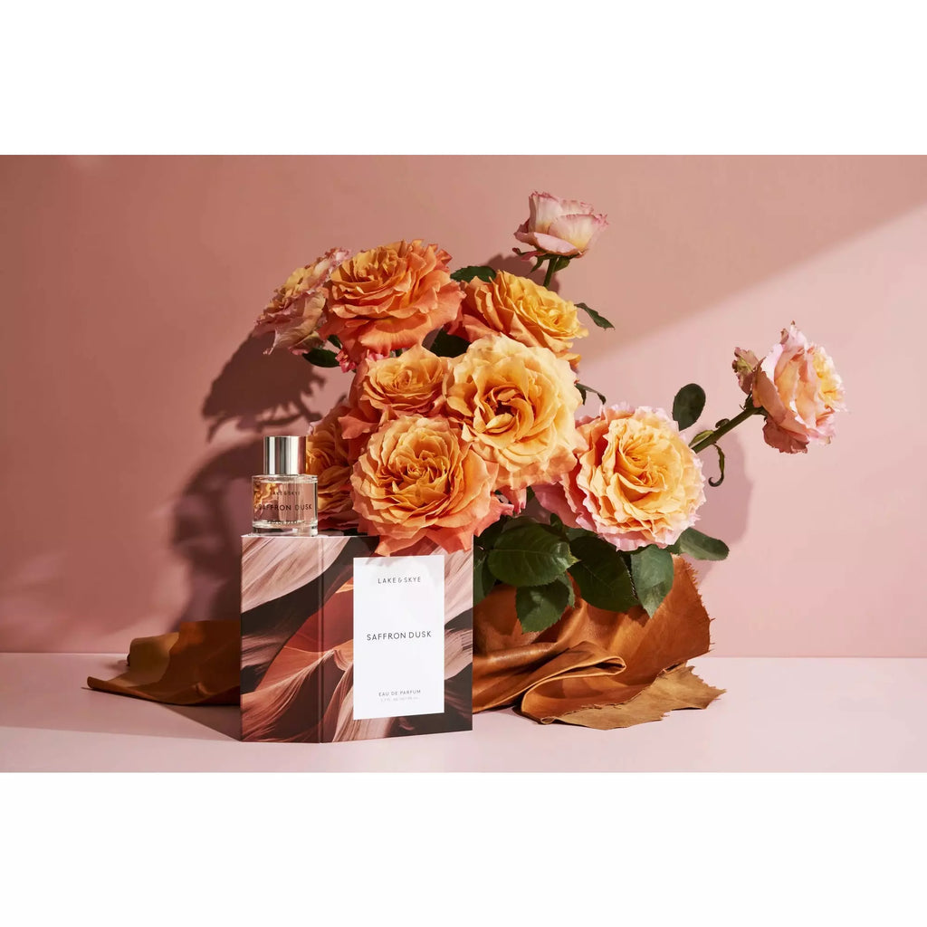 A bottle of perfume with its packaging placed beside a bouquet of orange roses against a pink background.