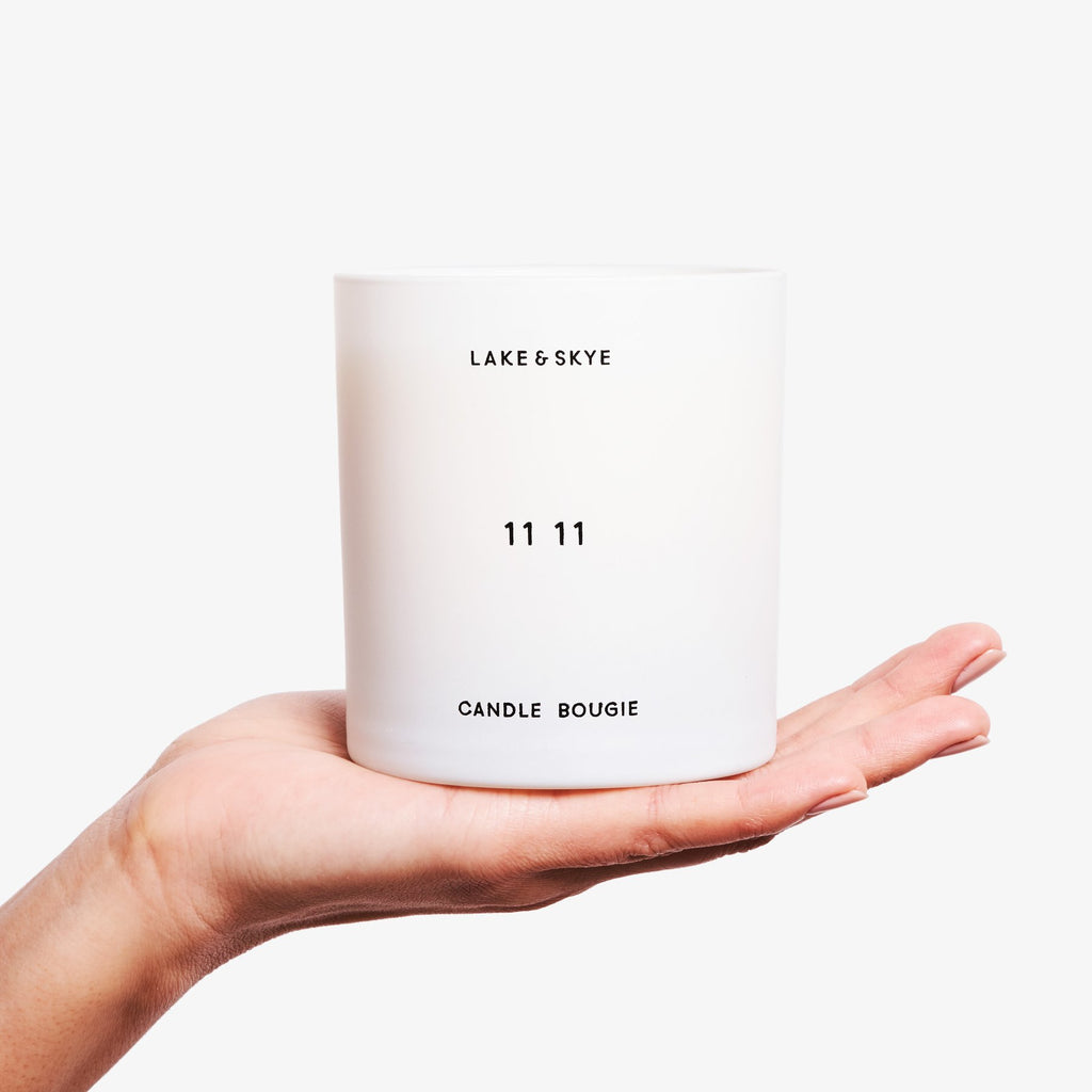 A hand holding a white "lake & skye 11 11" scented candle against a white background.
