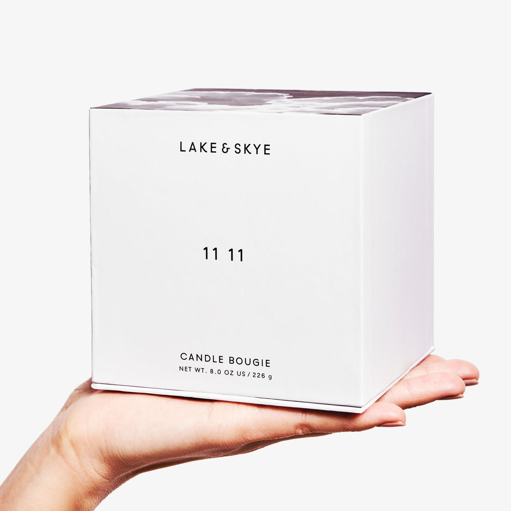 A hand holding a white box labeled "lake & skye 11 11 candle bougie" with a net weight indication.