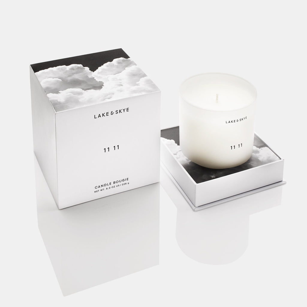 A white scented candle by lake & skye with the fragrance name "11 11" beside its packaging box, on a reflective surface.