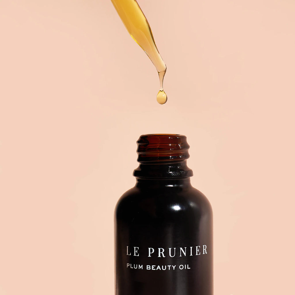 A dropper releases a single drop of plum beauty oil into a dark bottle against a pink background.