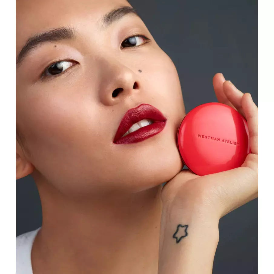 A person holds a red compact against their cheek with a focused gaze, displaying bold red lipstick and a visible star tattoo on the hand.