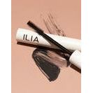 A close-up of a mascara brush and wand with the brand name ilia, against a pink background.