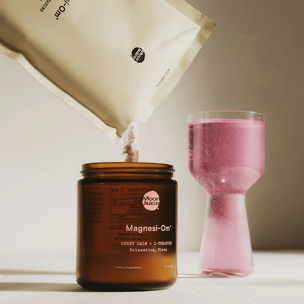 A powdered supplement is being poured from a beige pouch into a brown glass jar, with a pink glass filled with liquid nearby.