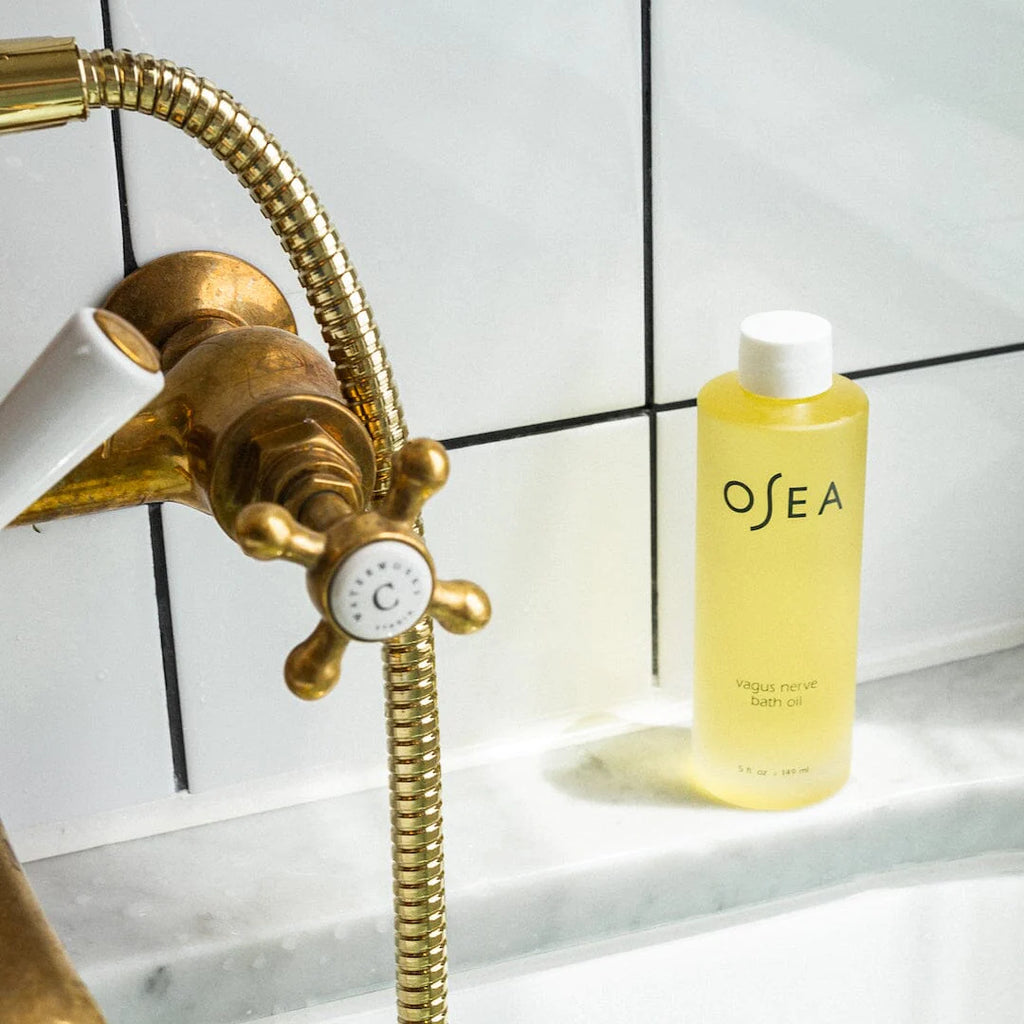 A bottle of osea bath oil beside a brass shower head and hose in a tiled bathroom.